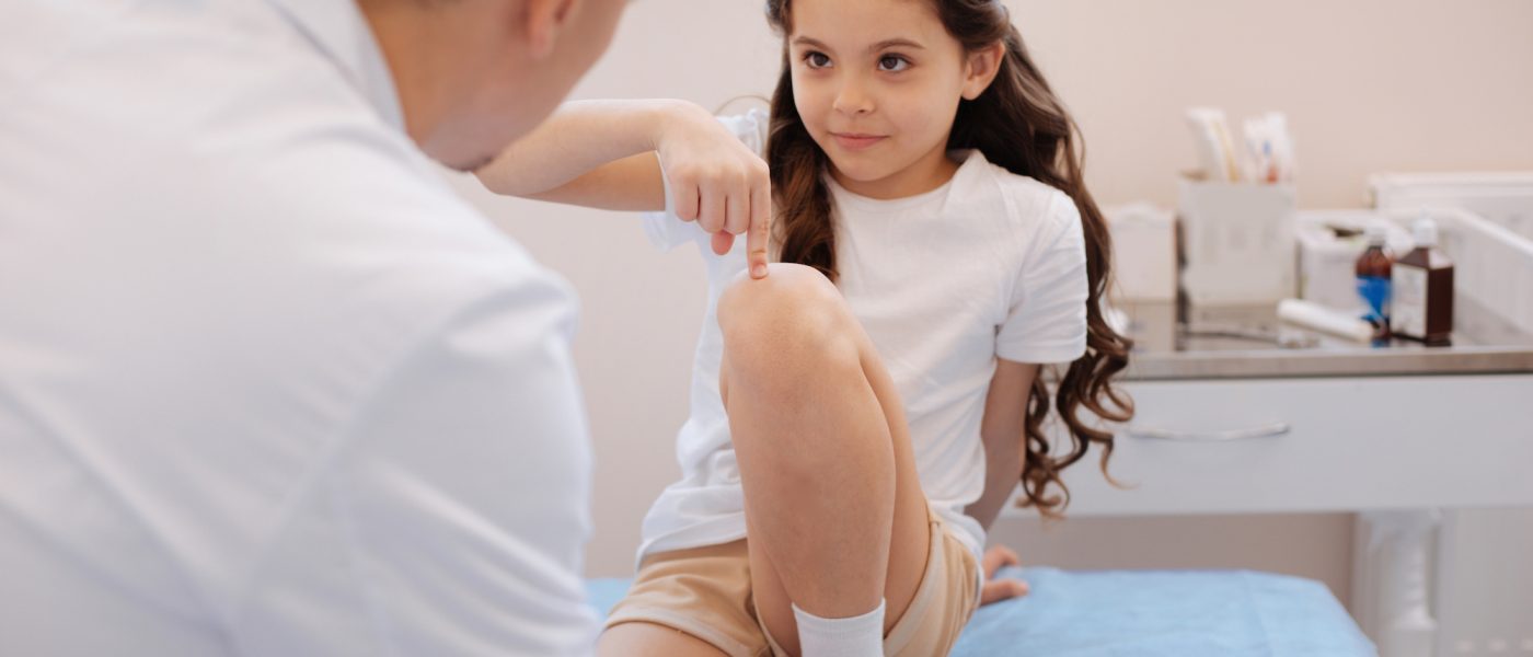 My knee hurts. Cute nice pleasant girl sitting on the medical bed and pointing at her knee while visiting a doctor