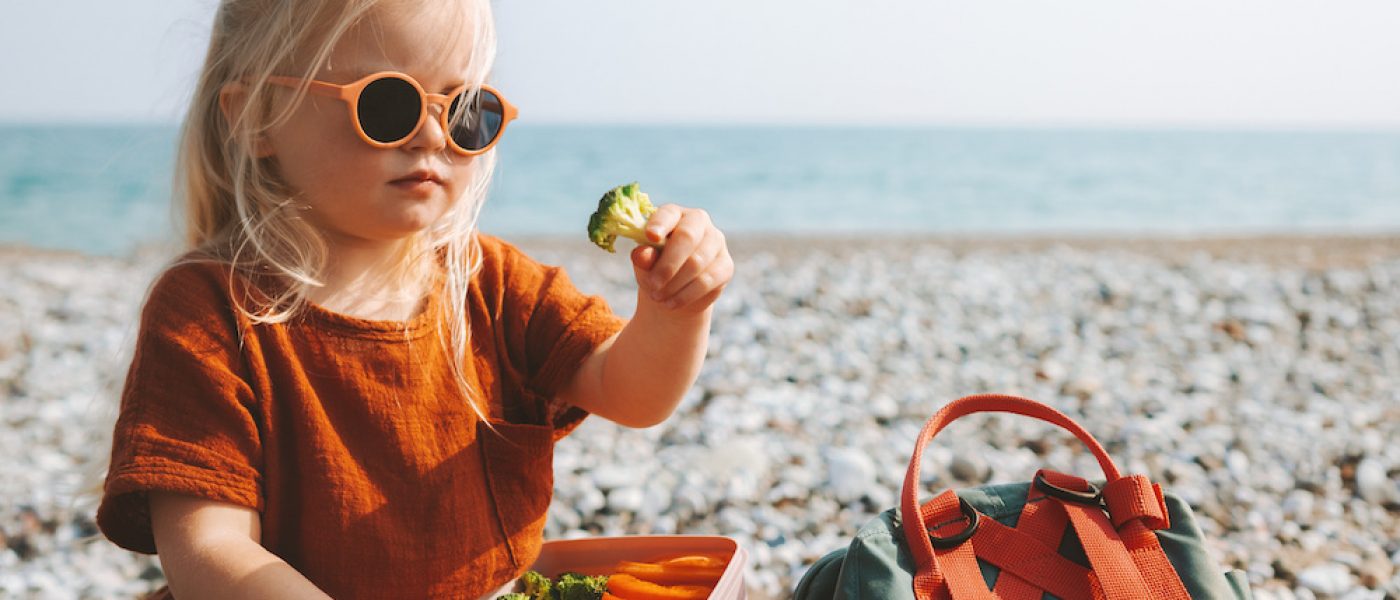 Child girl eating broccoli with lunch box picnic on beach vegan healthy food travel lifestyle outdoor summer vacations kid with backpack and lunchbox